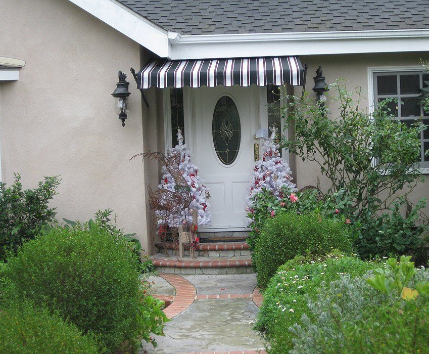 Front porch with white door, black-and-white awning, and garden path surrounded by lush shrubs and flowers