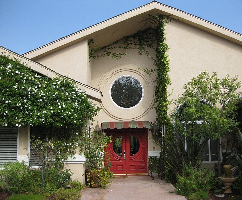 Beige house with circular window above red double doors, framed by green shrubs and vines.