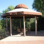 A gazebo with a copper-colored domed roof and scalloped trim on a paved patio, surrounded by trees.