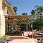 Patio with pergola beside a beige house, brick steps, and palm trees in bright sunlight