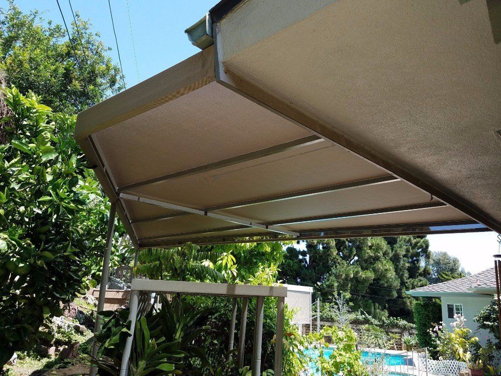 Beige patio awning over a backyard with trees and a pool in bright sunlight