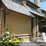 Tan awning shading a beige house exterior with stone trim and flowering garden in front
