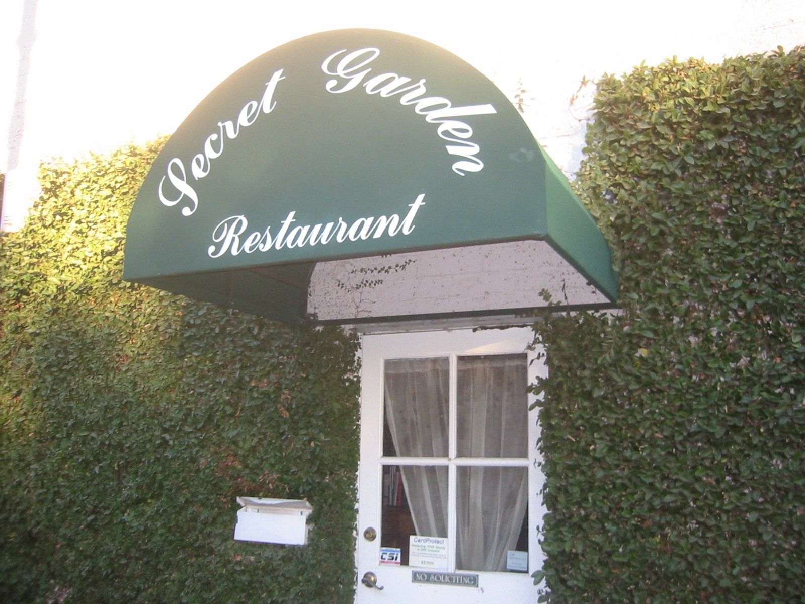Restaurant entrance with green awning reading “Secret Garden,” set against a hedge wall