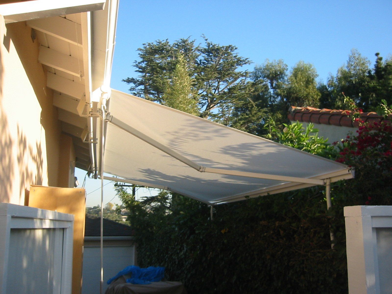 Backyard patio with a white shade sail stretched from a house to posts, under a blue sky.