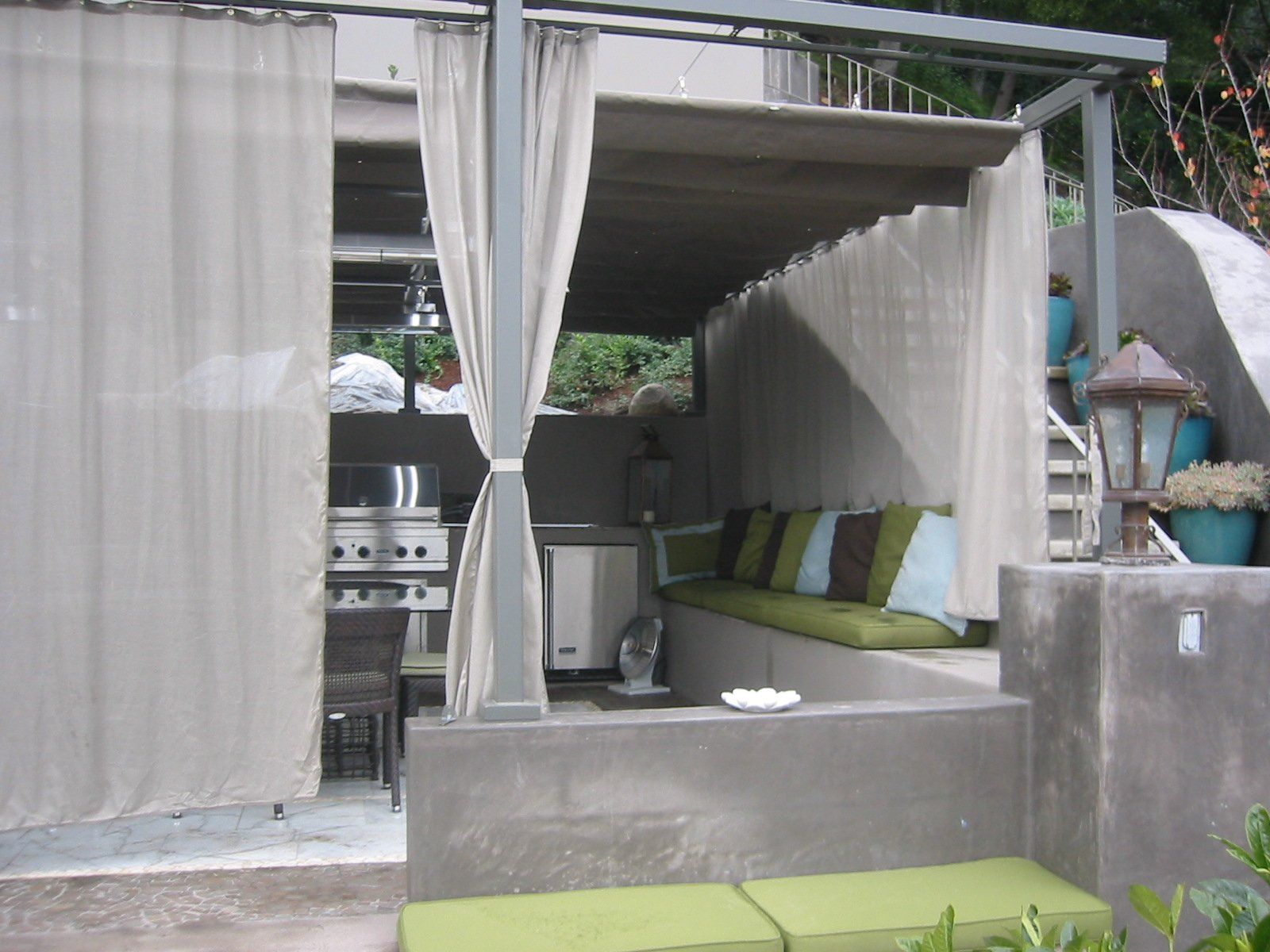 Outdoor cabana with sheer curtains, green-cushioned bench, and dining table beside a poolside wall.