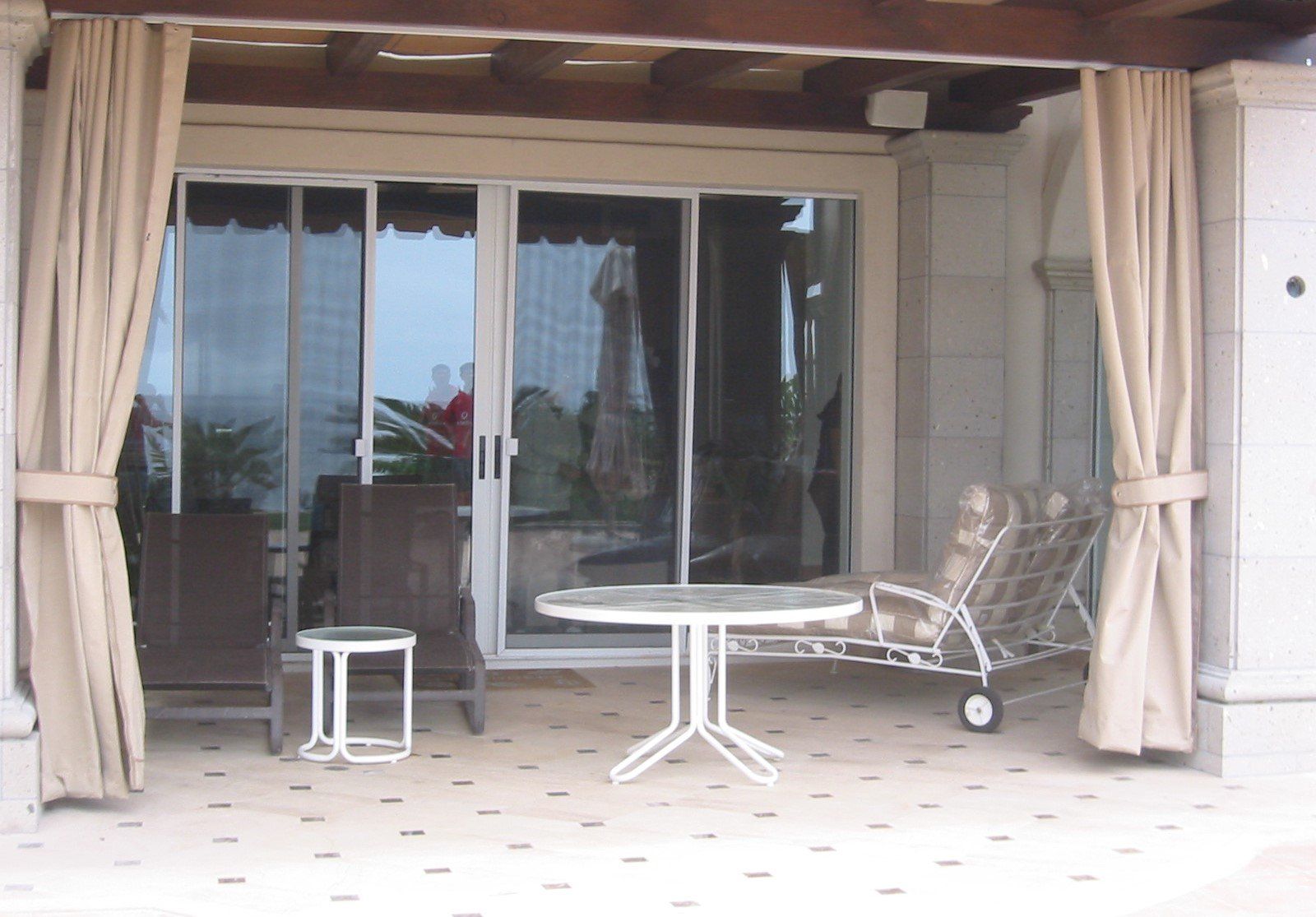 Covered patio with sliding glass doors, beige curtains, white table, small stool, and lounge chairs.