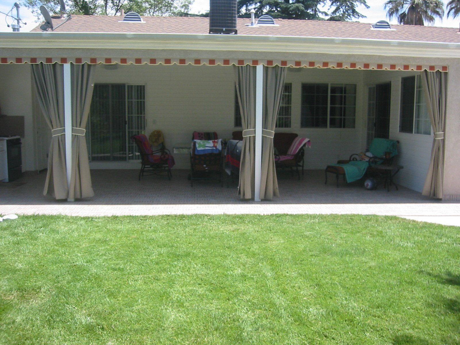 Covered patio with curtains, outdoor chairs, and a green lawn in front of a house