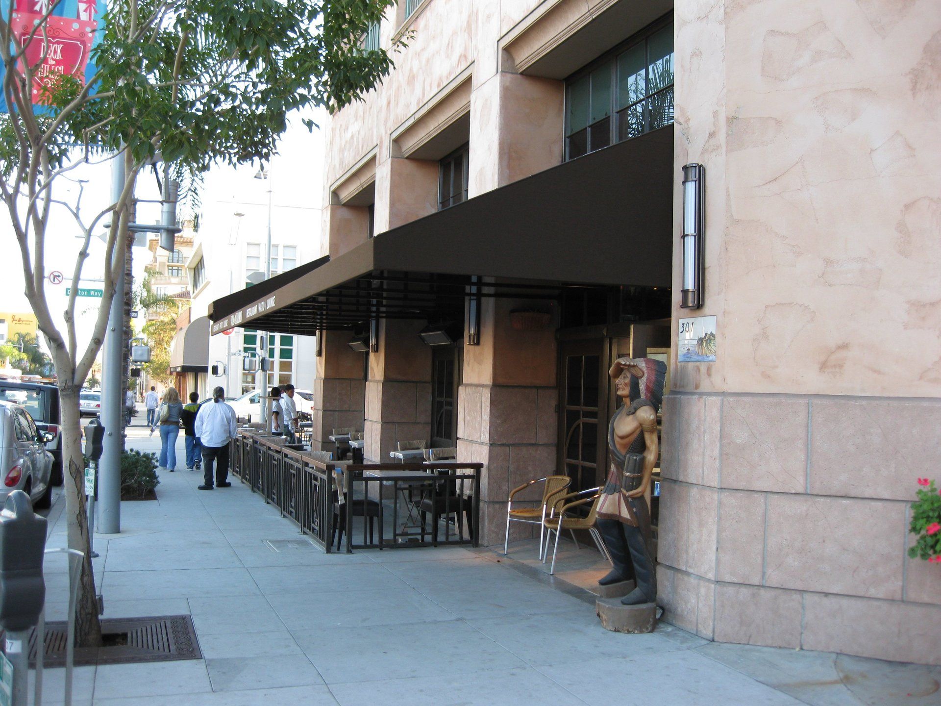 Sidewalk café with black awning, outdoor tables and chairs beside a stone building on a sunny street.
