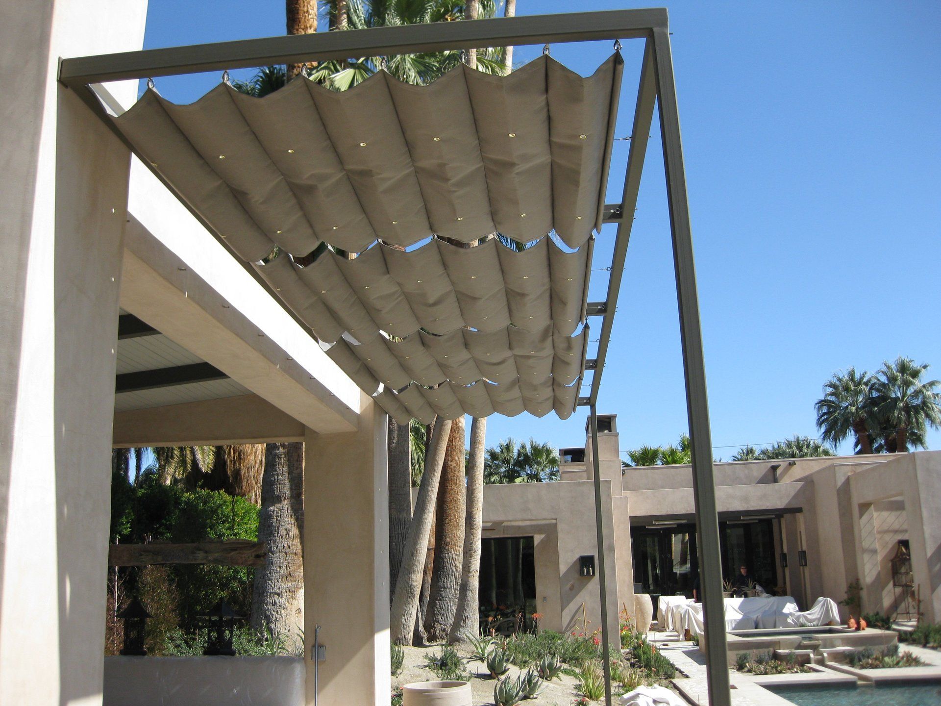 Modern patio with a beige retractable canopy beside a resort-style pool and palm trees