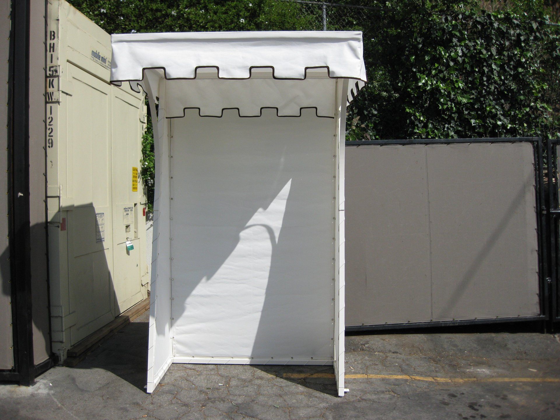 White canopy booth or kiosk against a wall, casting a shadow in a sunlit outdoor area.