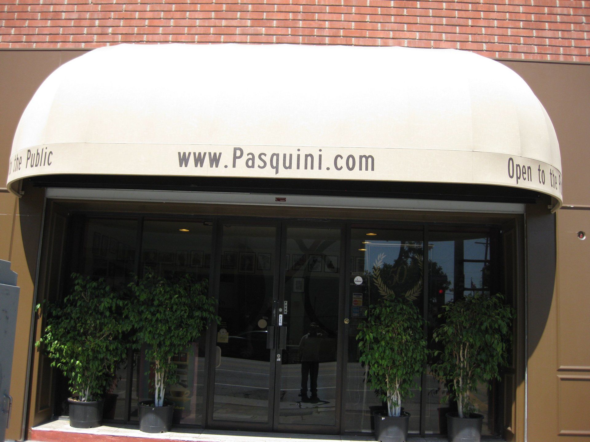 Storefront with white awning and black glass entrance, flanked by potted trees under a brick facade.