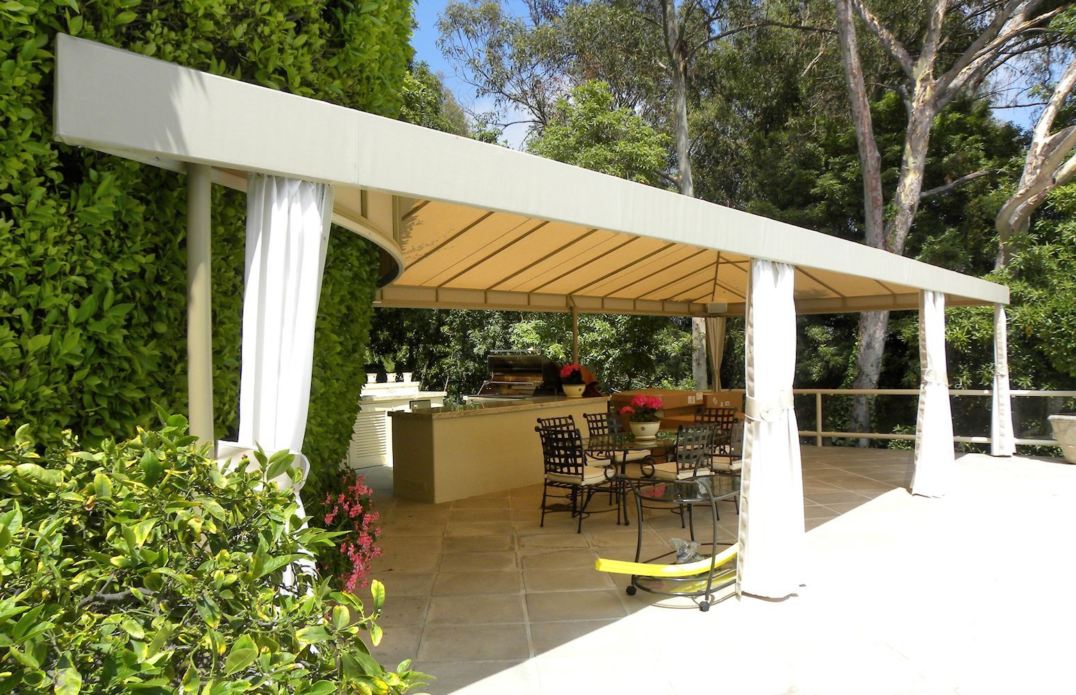 Outdoor patio with white pergola, curtains, and a tiled seating area surrounded by lush greenery