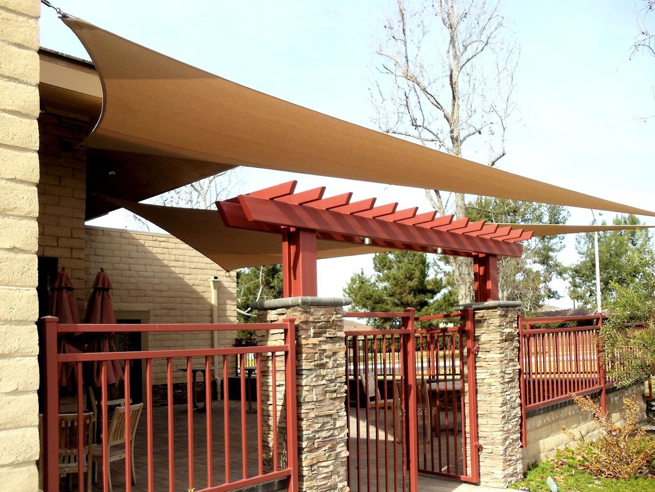 Patio entrance with red metal pergola, tan shade sails, and stone columns behind a red fence