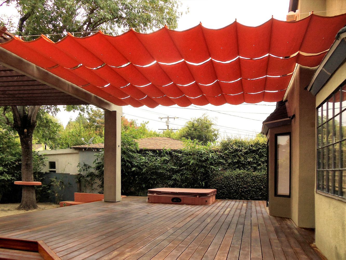 Patio with red shade sails over a brick floor, trees, and a bench beside a building.