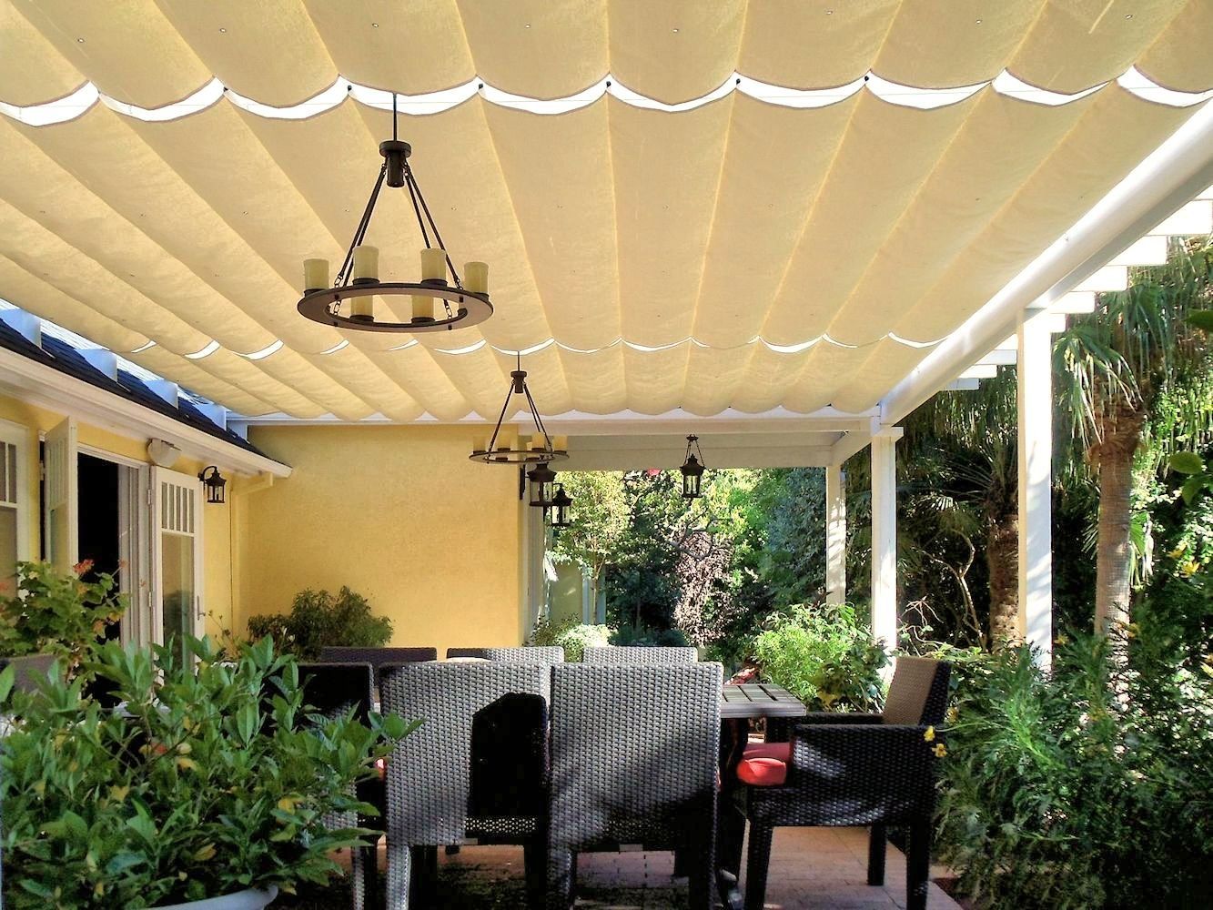 Covered patio with hanging lights, wicker seating, and lush green plants beside a house.