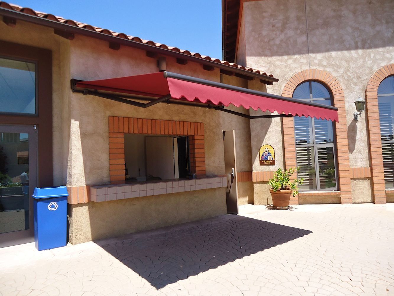Outdoor service window with red awning on a stucco building, next to a blue trash bin.