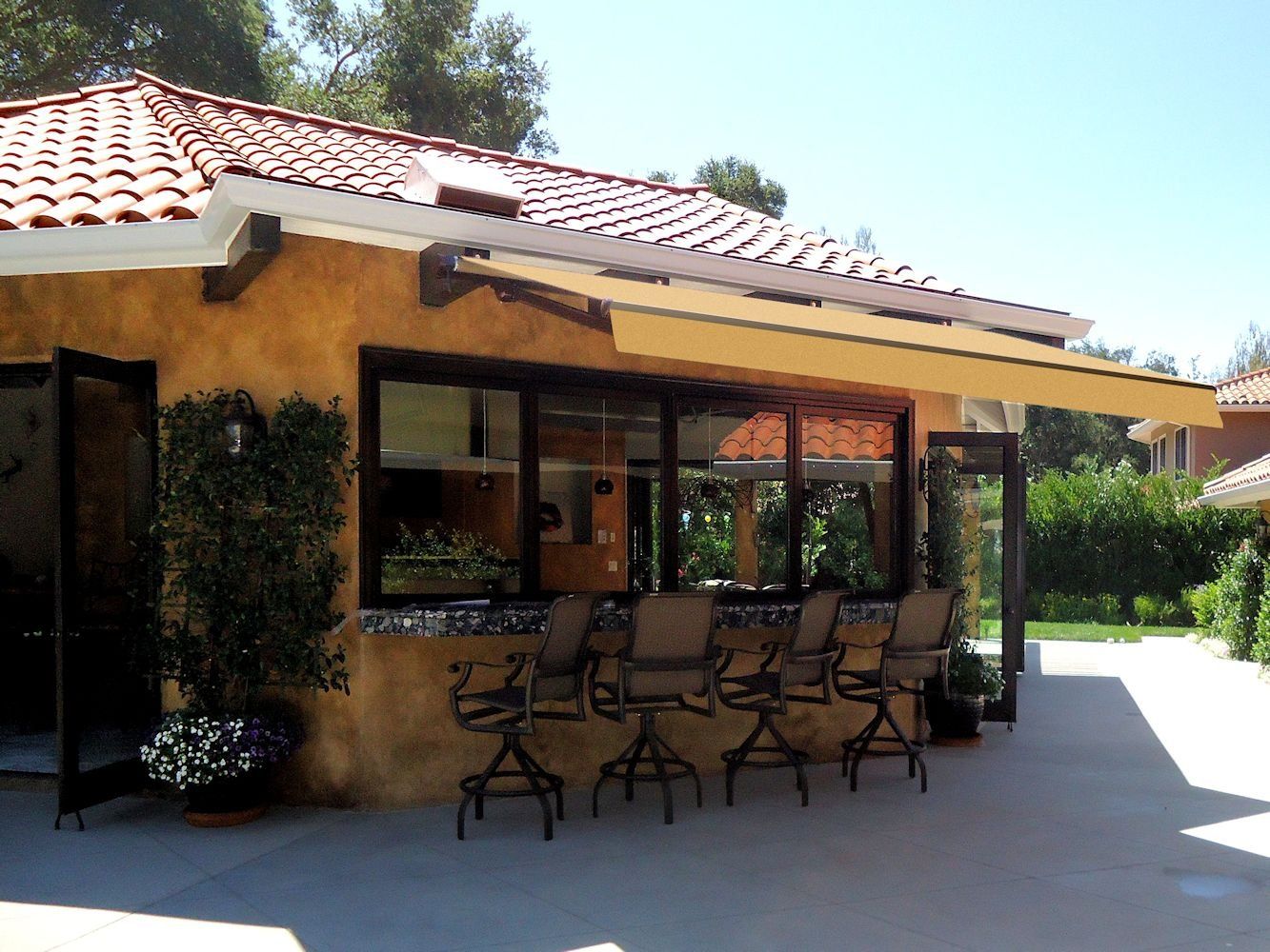 Outdoor patio with black metal chairs beside a stucco house with a red tile roof and awning