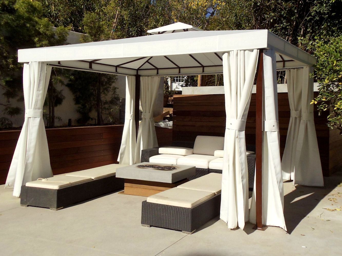 White canopy daybed with curtains and tan cushions on a patio, with bench seating on each side