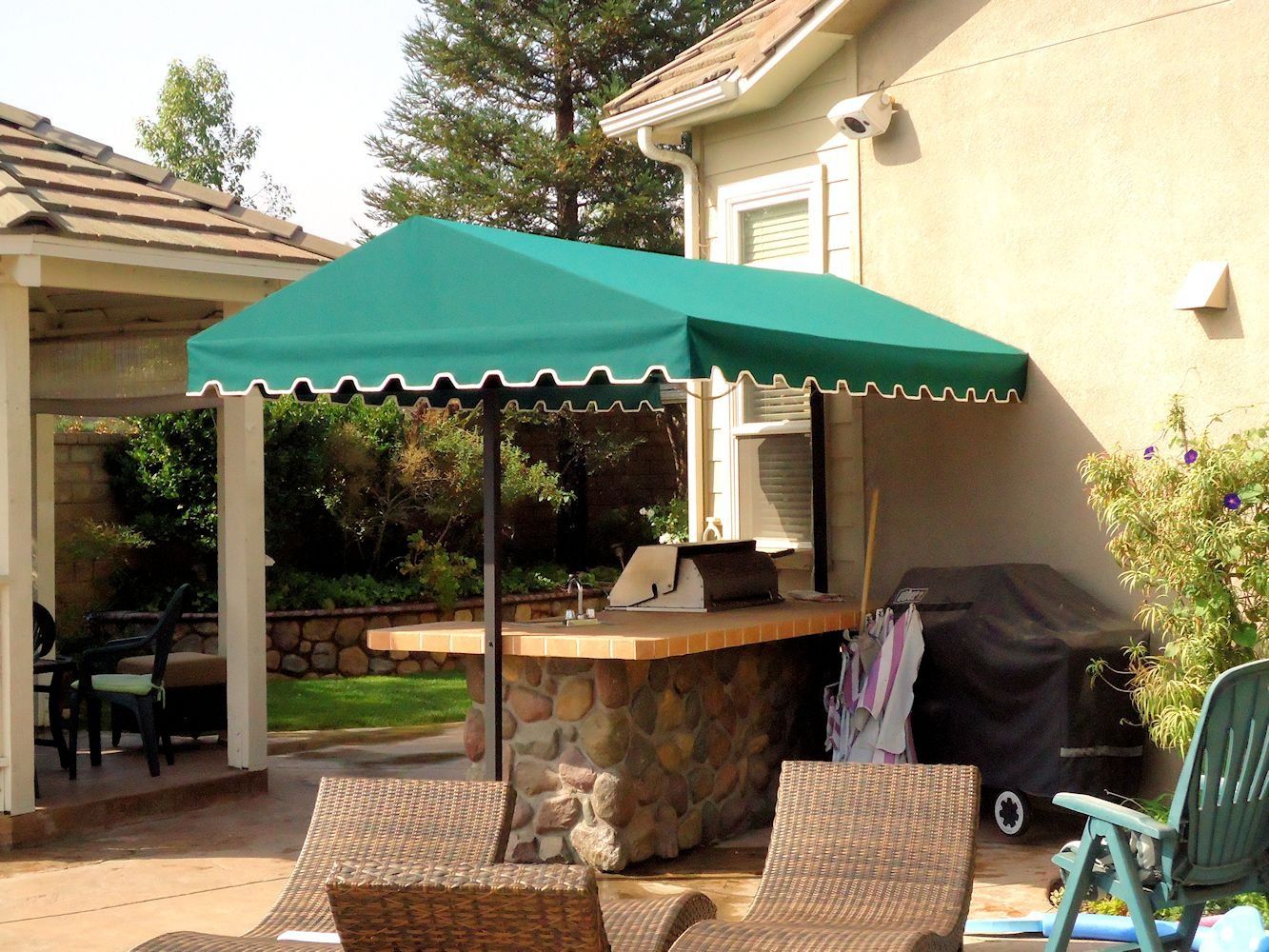 Backyard patio with green canopy over stone bar, lounge chairs, and a grill beside a house