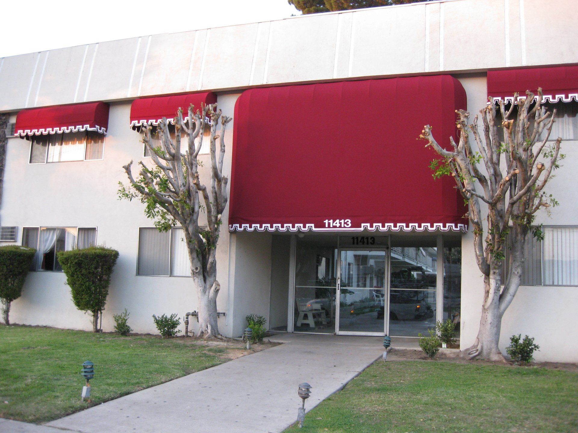 Building entrance with red awnings, glass doors, and trimmed trees along a sidewalk and lawn