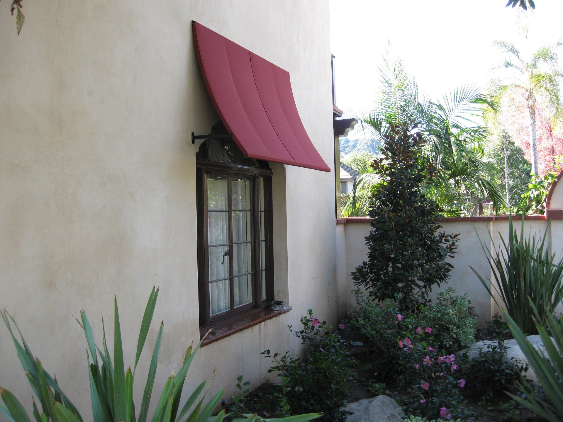 Stucco house wall with a window and red awning beside a small garden and shrubs