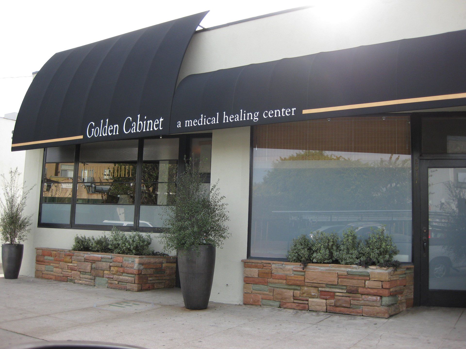 Storefront with black awning, large windows, and potted plants outside a sidewalk.