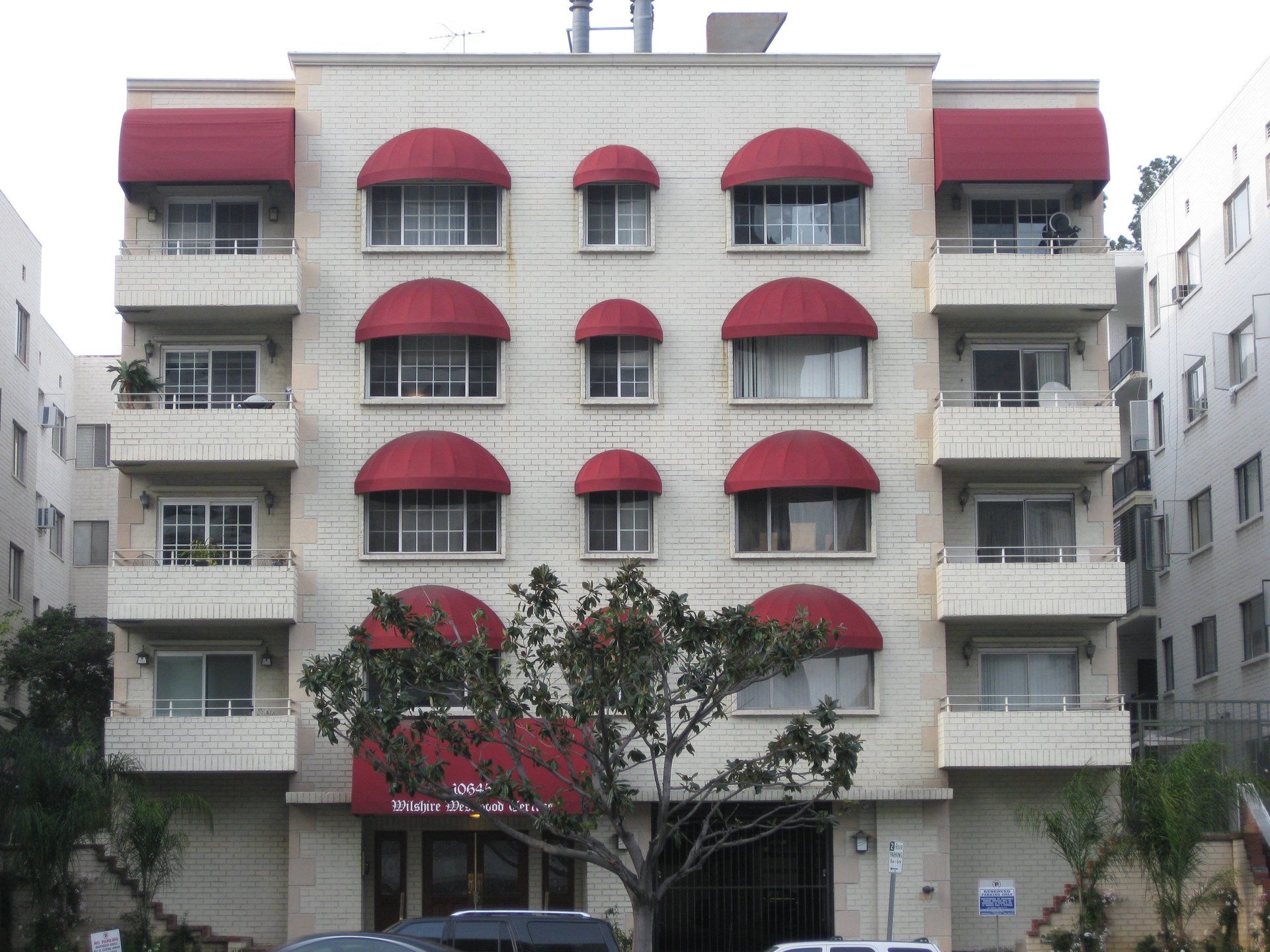 Apartment building facade with red awnings and balconies, partially obscured by trees