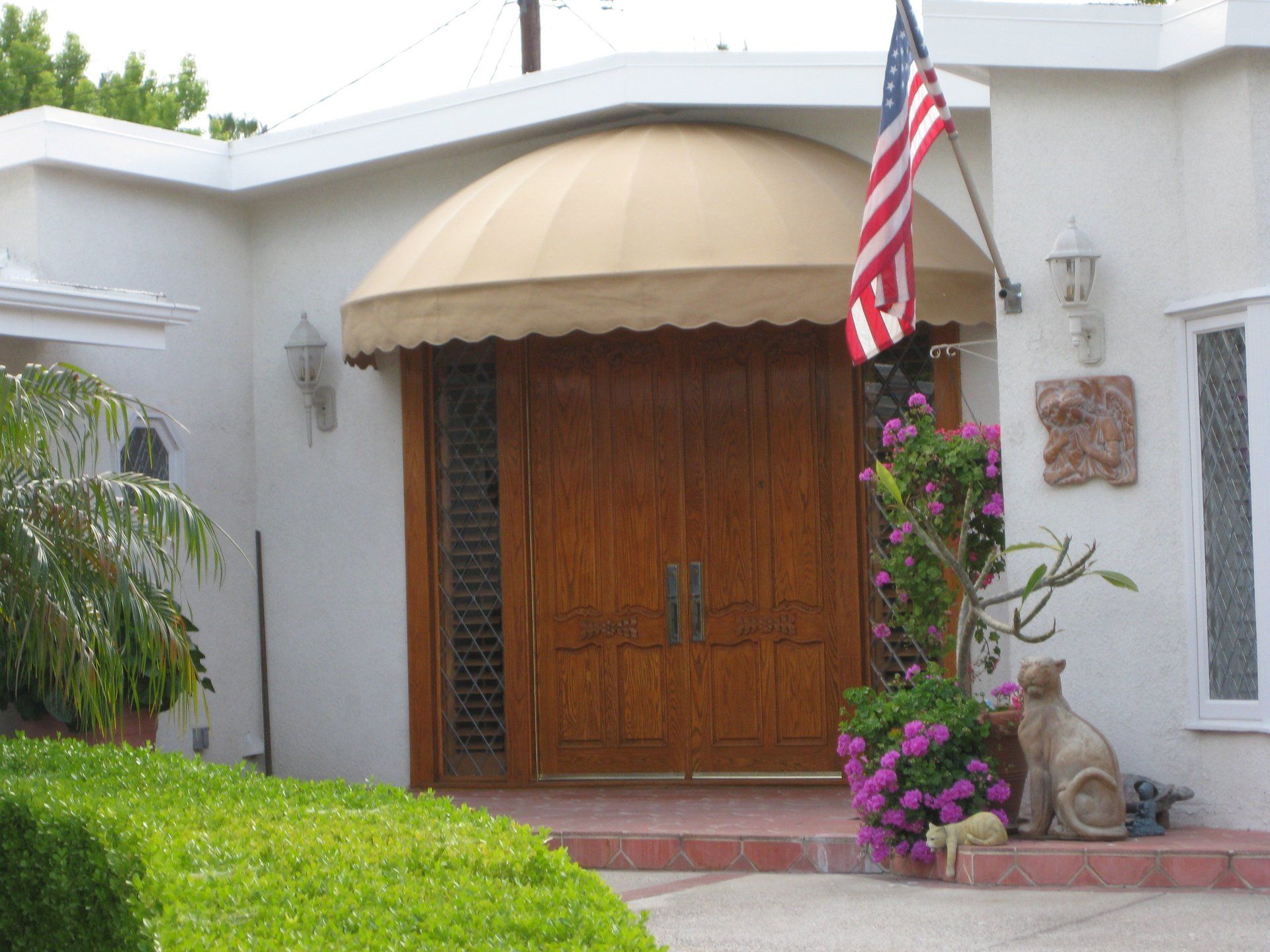 Front entrance with wooden double doors, beige awning, American flag, and flowers beside a white stucco wall