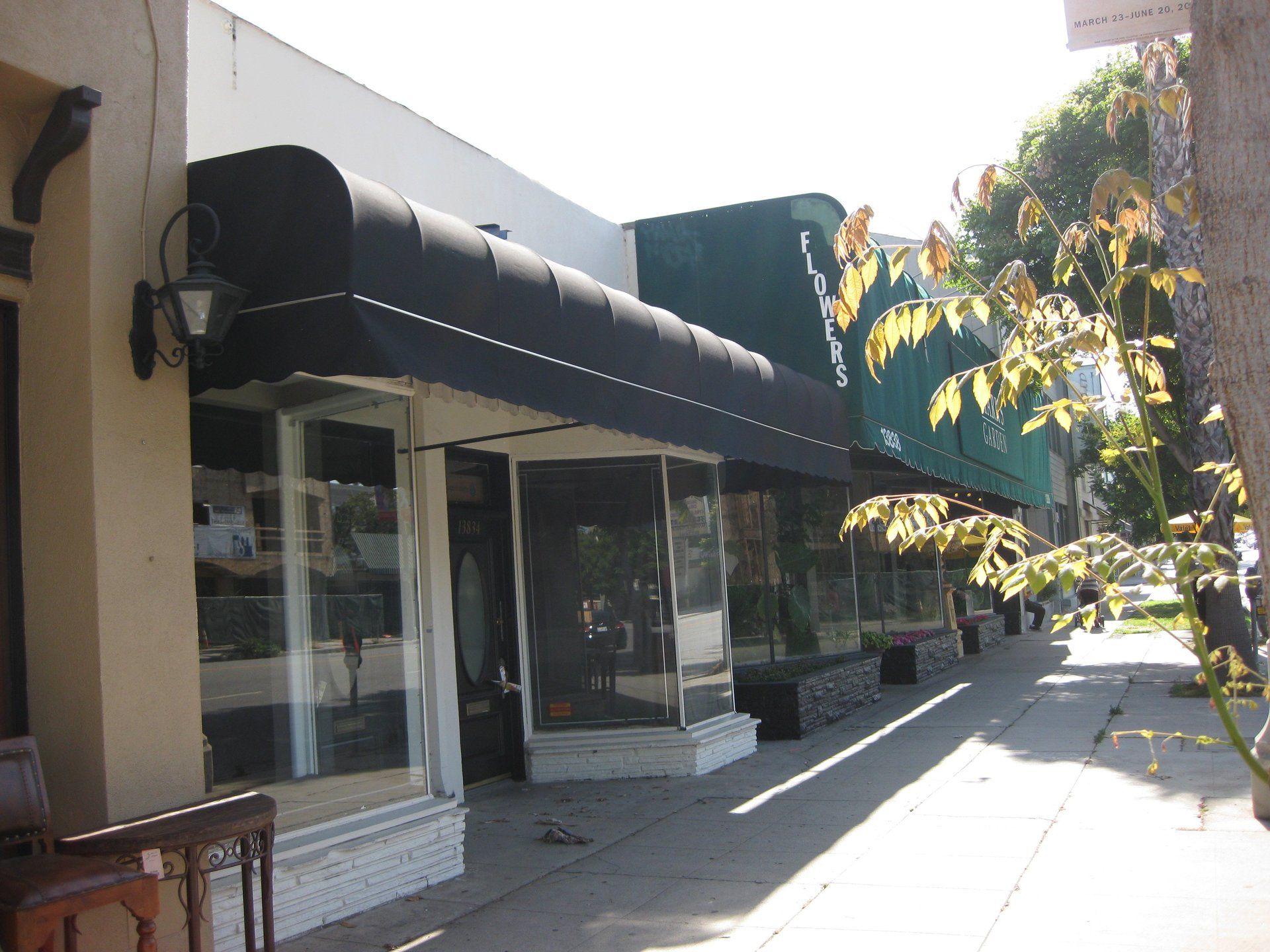 Sidewalk storefronts with black awnings, glass windows, and trees casting shade on a sunny street.