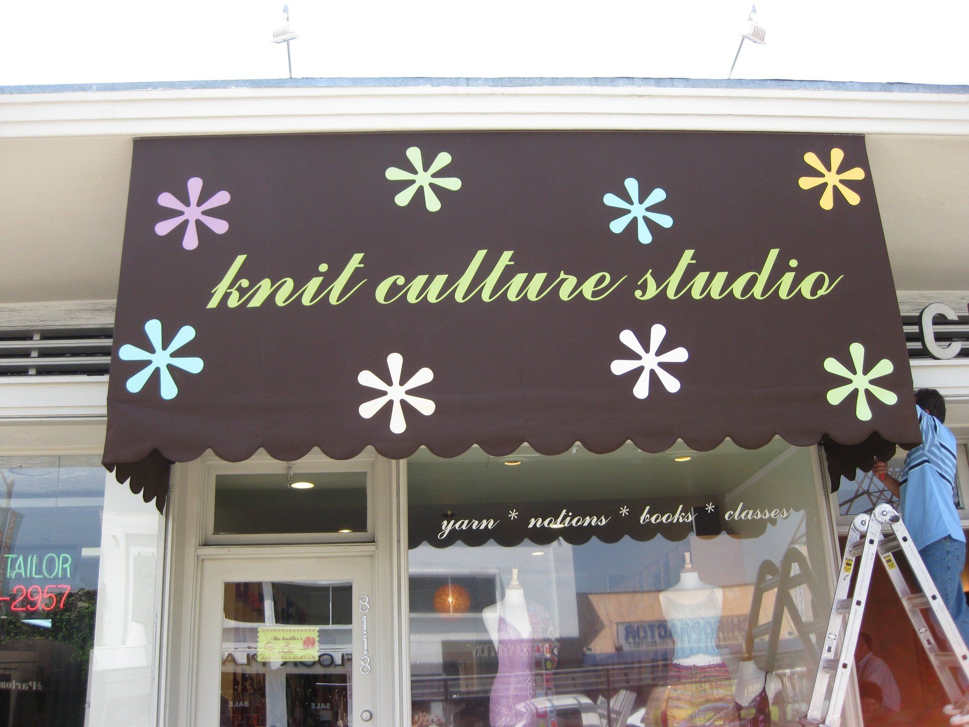 Storefront sign for “knit culture studio” on a brown awning with pastel starburst flowers