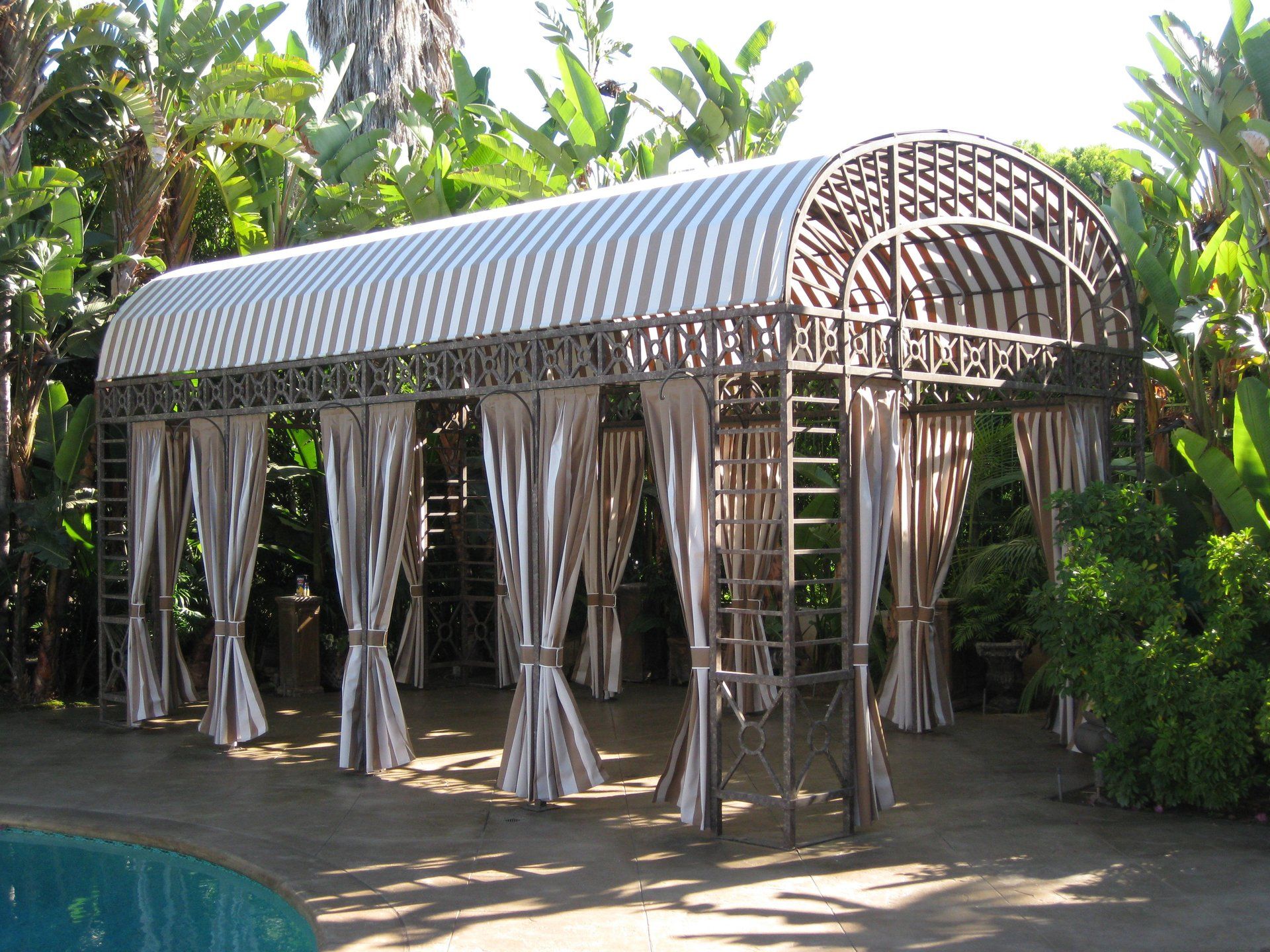 Poolside gazebo with striped curtains and a curved metal roof, surrounded by tropical plants