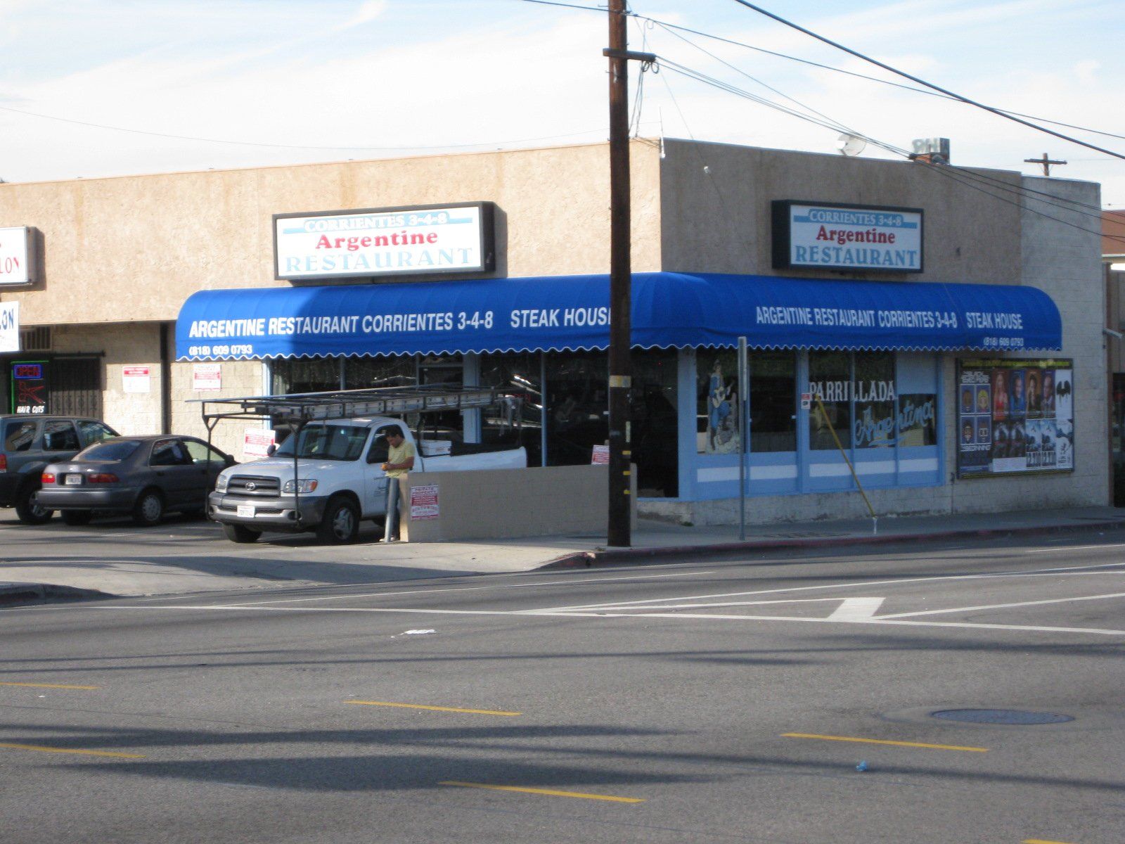 Street-corner auto repair shop with blue awnings, parked cars, and a beige storefront.