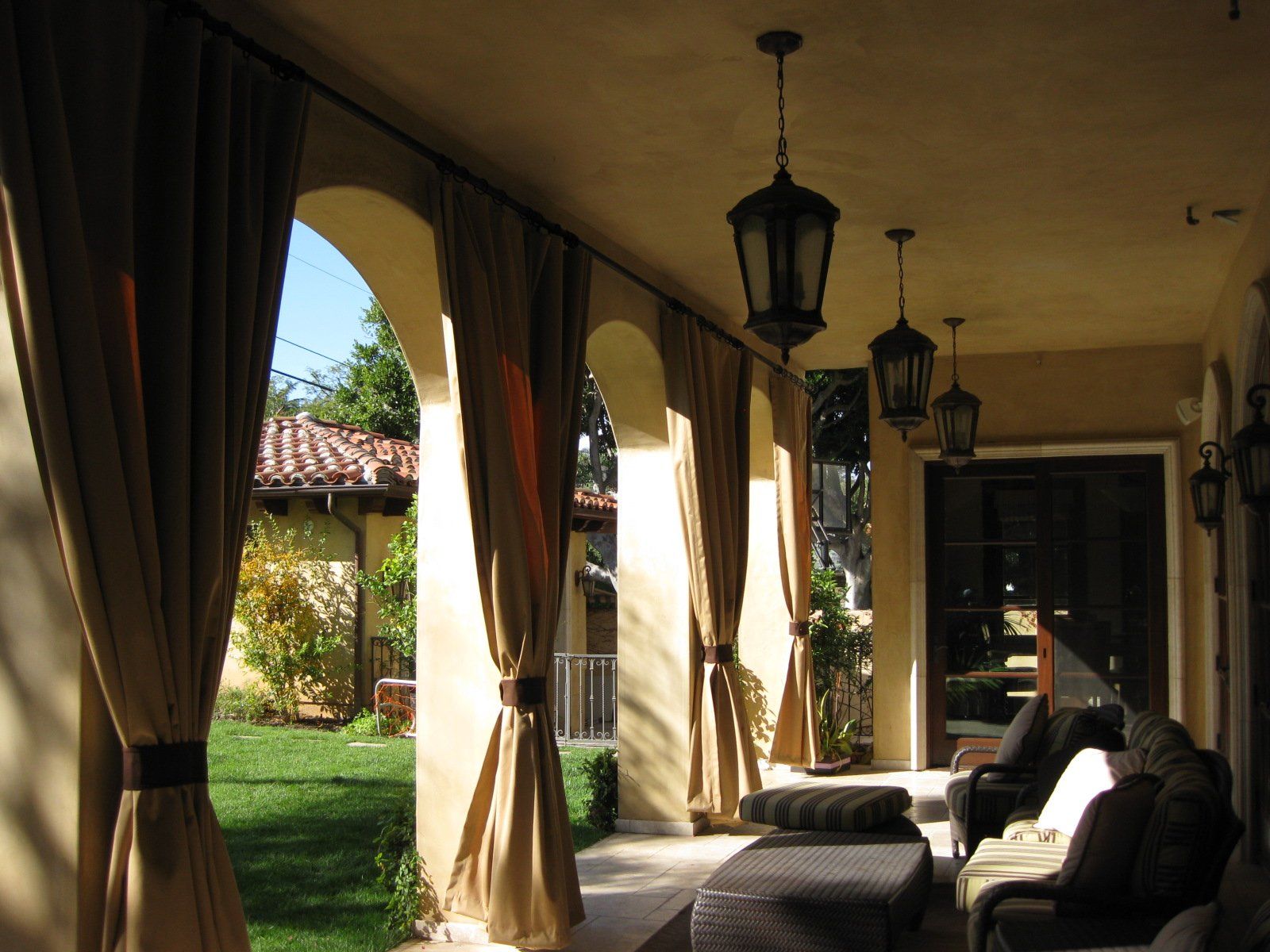Covered patio with hanging lanterns, curtains, and cushioned lounge chairs overlooking a grassy yard.