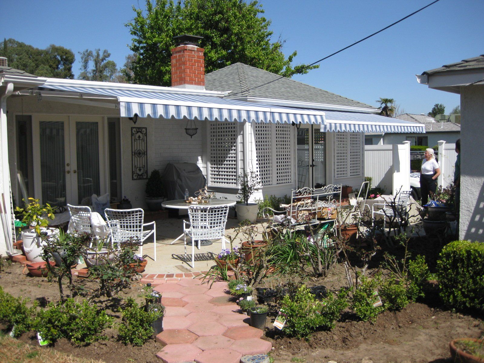 Front yard garden with a white house, patio chairs, and a brick walkway under a striped awning.