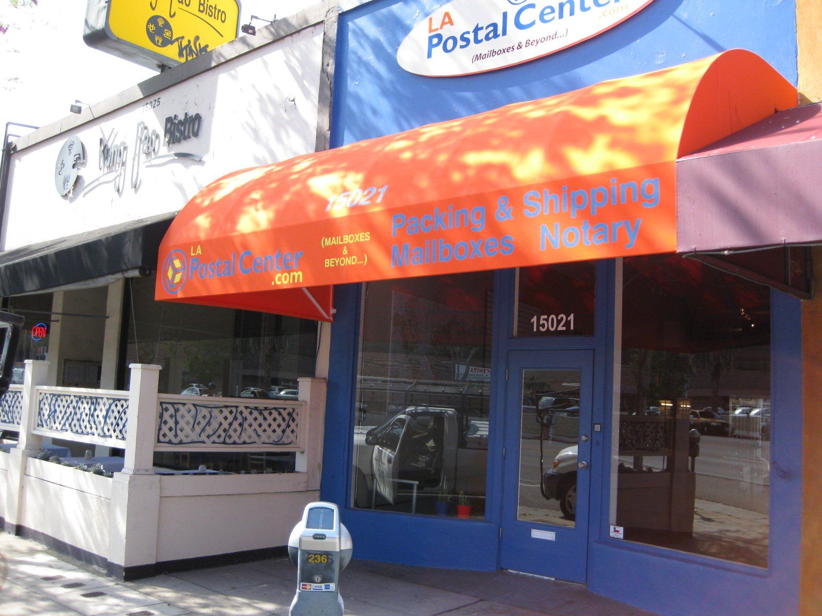 Storefront with orange awning for a Postal Center, featuring blue doors and a sidewalk parking meter.