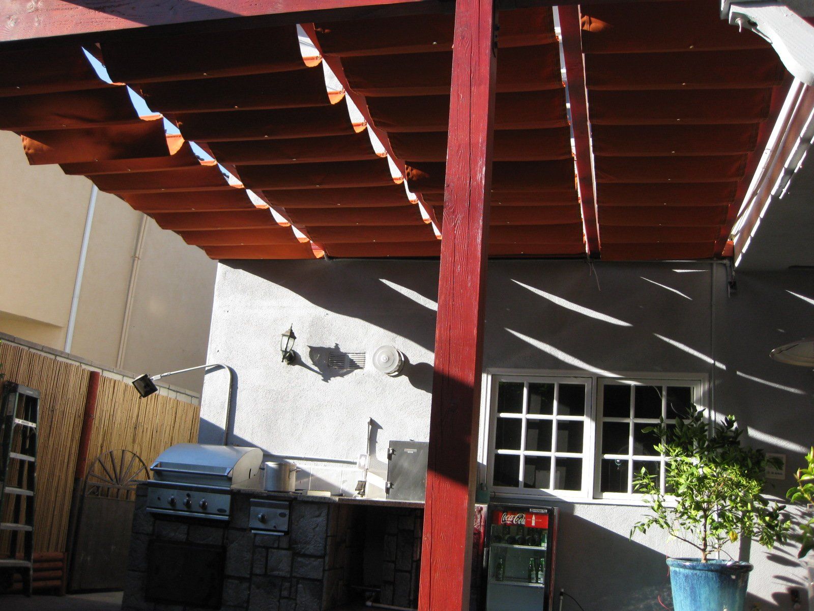 Backyard patio with red pergola beams, trash bins, a windowed wall, and potted plants in sunlight
