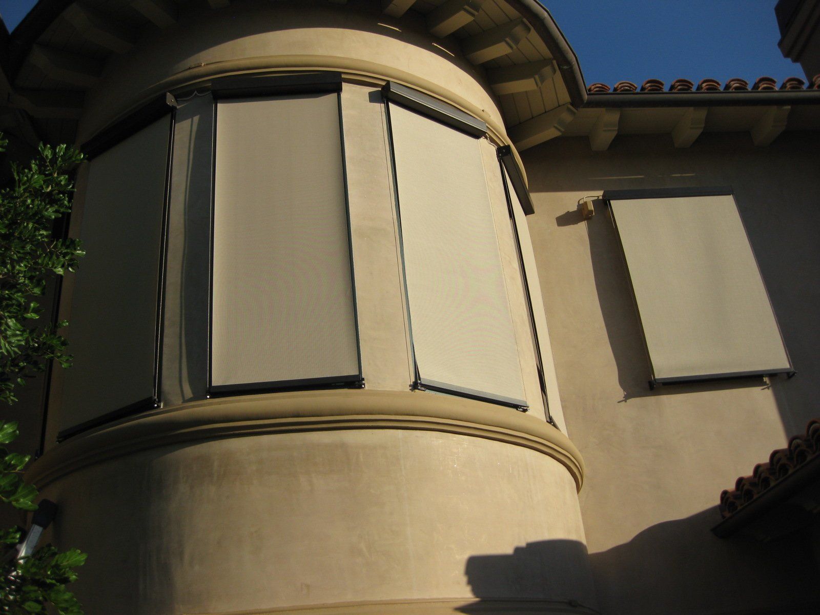 Tan stucco tower with curved bay window and closed shutters beside a tiled roof under blue sky