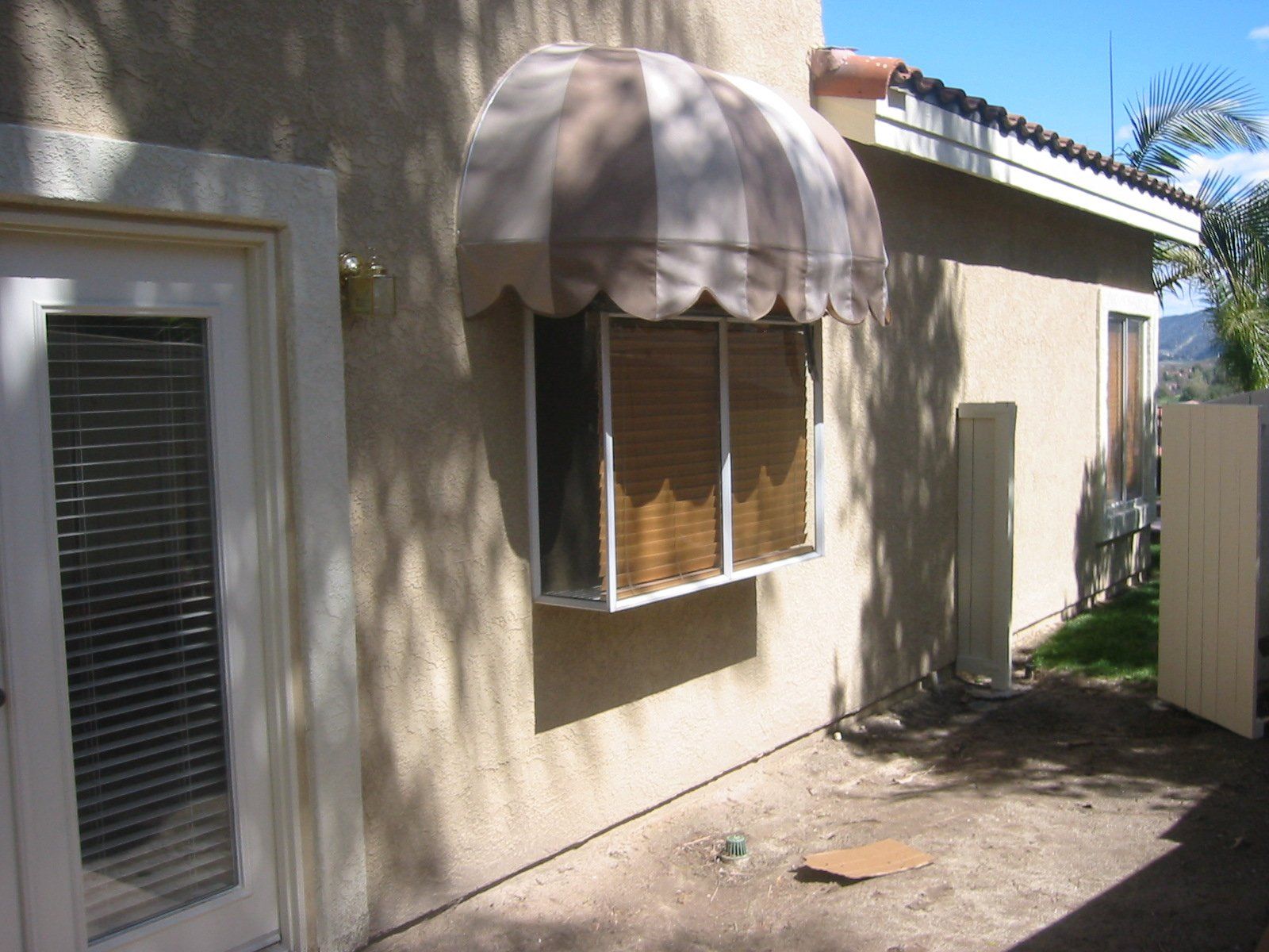 Back patio with beige stucco wall, striped awning over window, and a sliding glass door on the left.