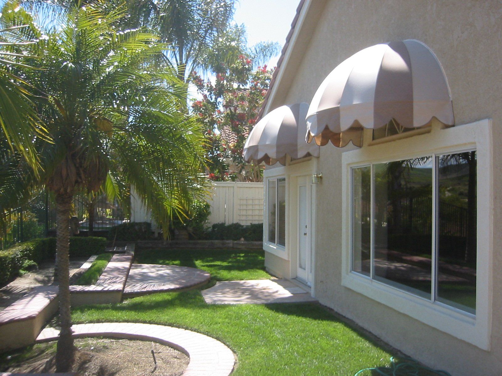 Sunny backyard patio with palm trees, lawn, and large windows under beige awnings