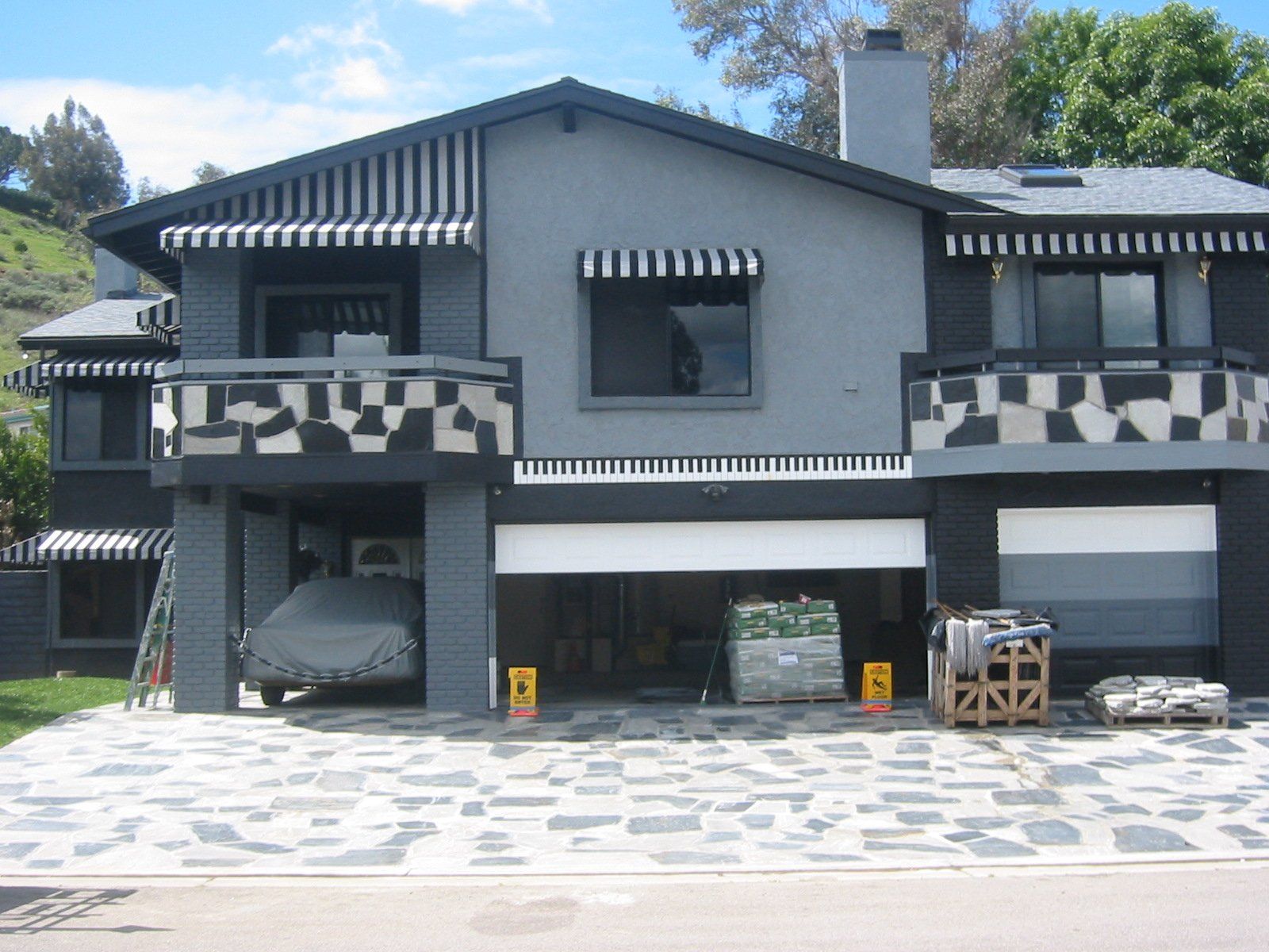 Two-story gray house with striped awnings, garage doors, and a stone driveway in daylight