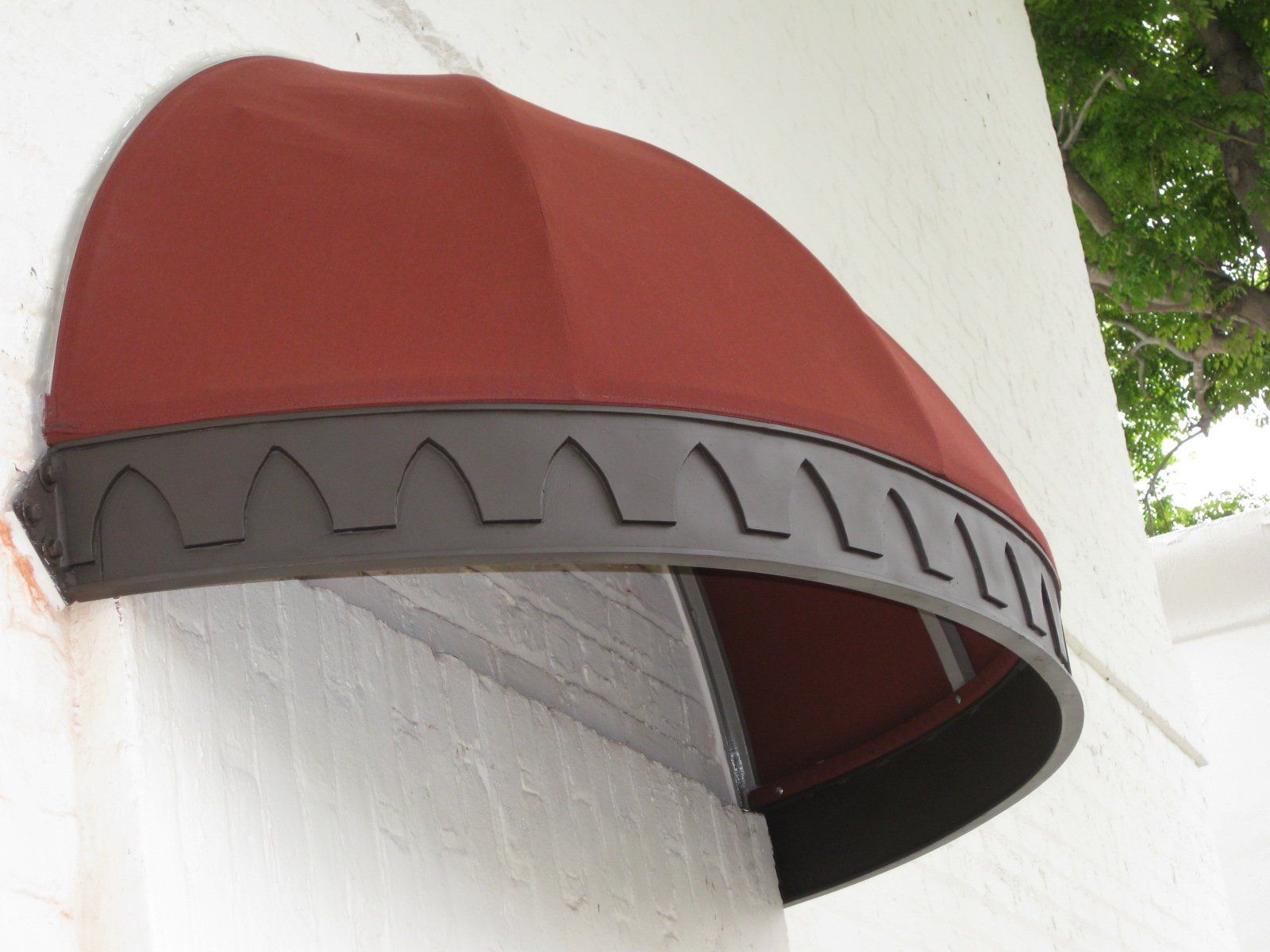 Red-brown awning with gray trim mounted on a white wall near greenery.