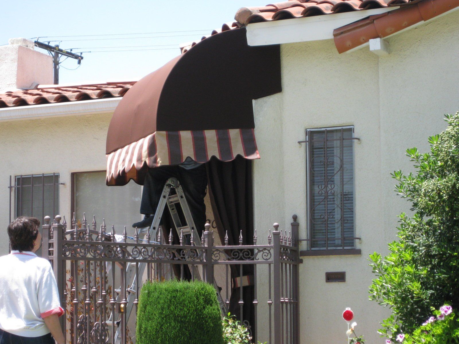 Person approaching a house with a brown awning and metal fence, viewed from the sidewalk