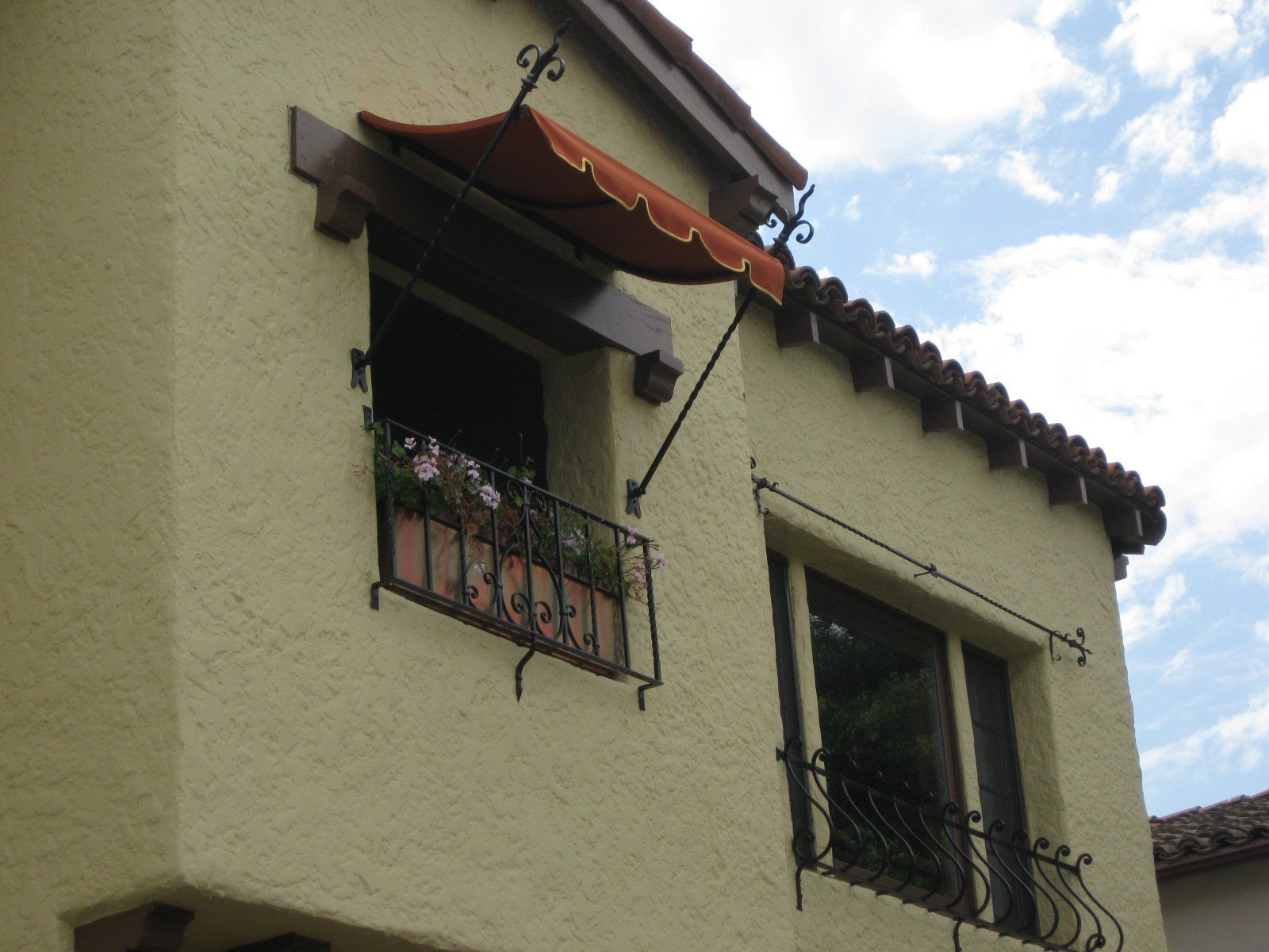 White stucco house facade with two wrought-iron balconies and a red awning under tiled roof eaves