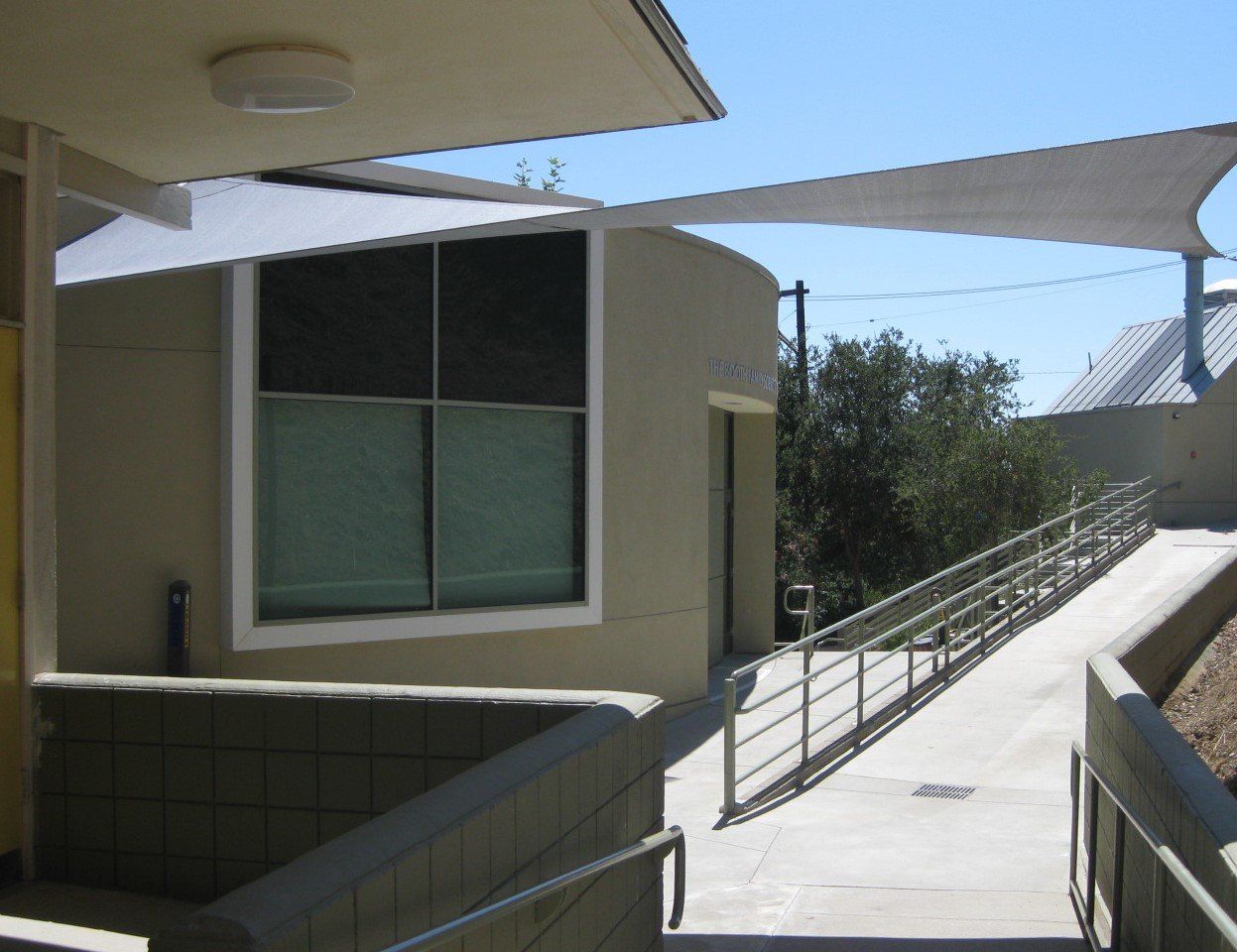 Modern building walkway with ramps, railings, and shade sails in bright sunlight