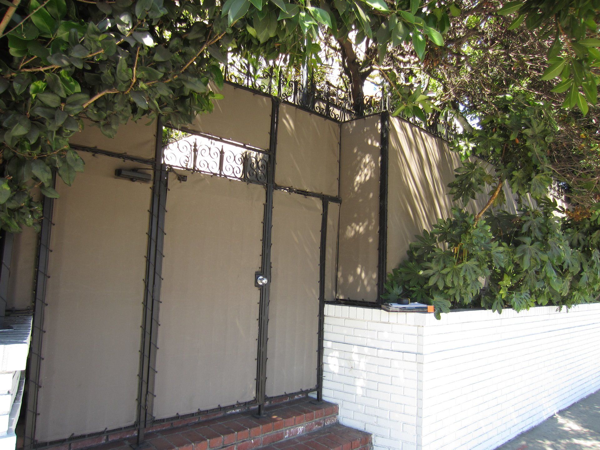 Gated entrance beside a white brick wall, partly covered by tree branches.