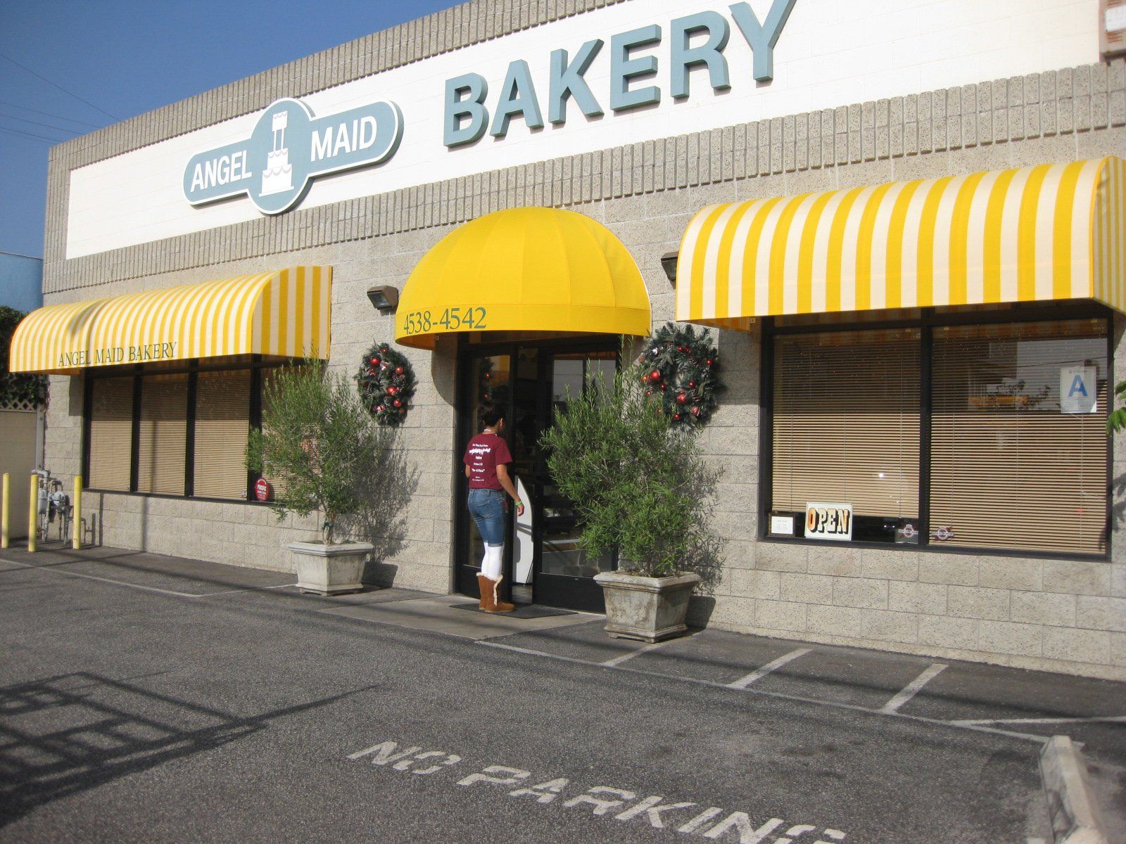 Bakery storefront with yellow awnings and a person at the entrance; no parking painted on the lot.