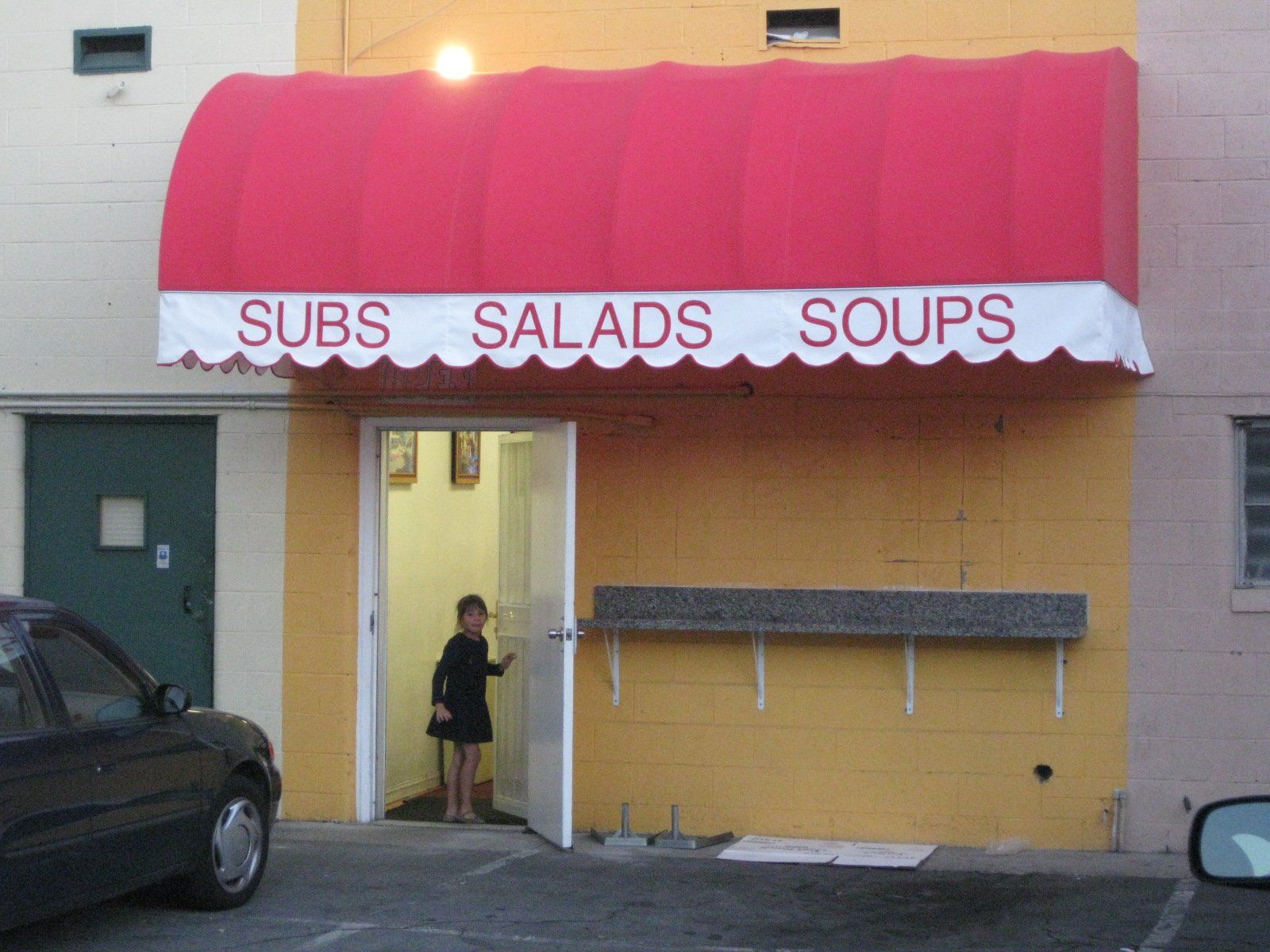 Person standing in doorway of yellow storefront with red awning labeled “Subs Salads Soups”