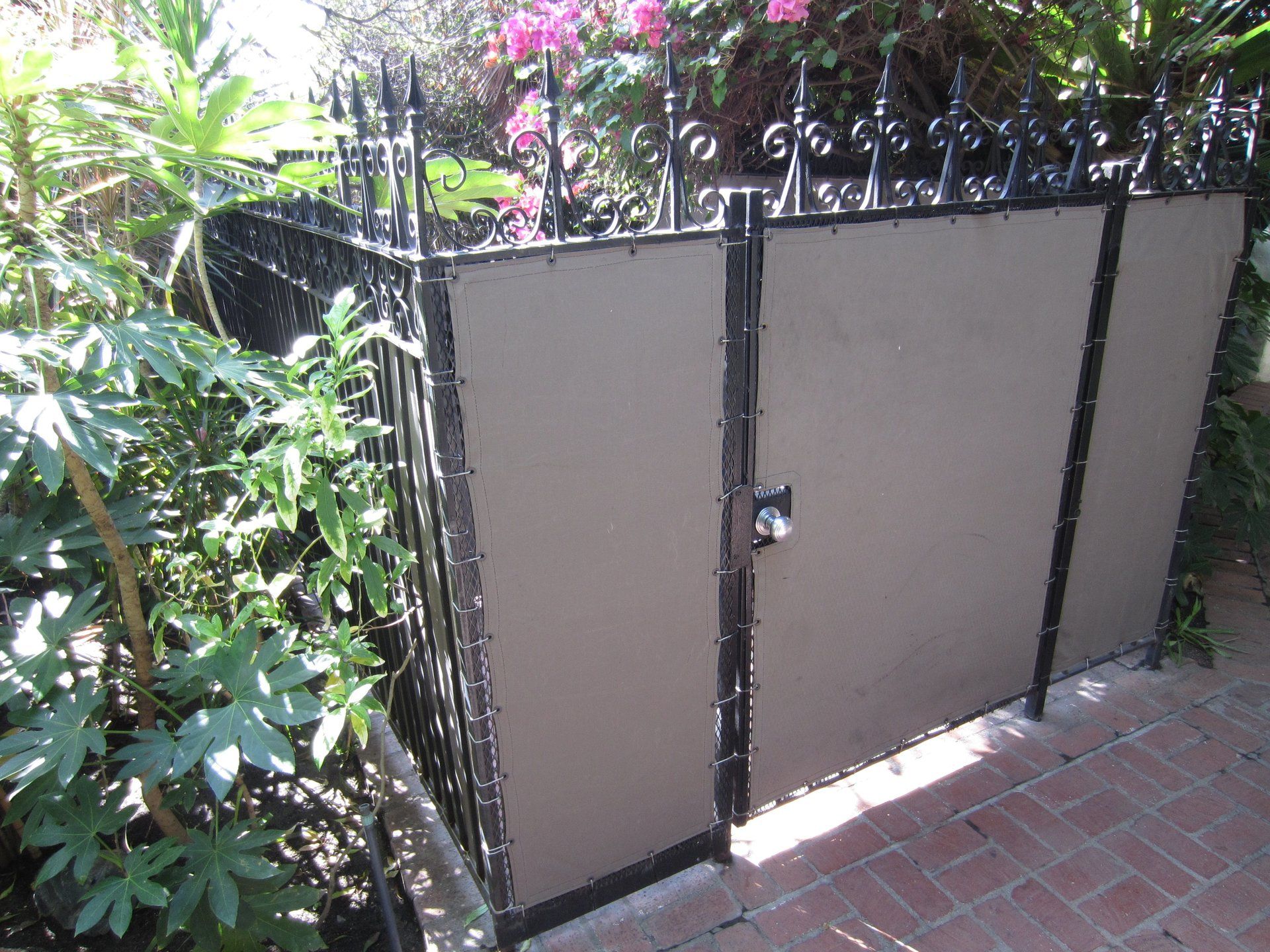 Closed beige metal gate beside lush green plants and a brick walkway