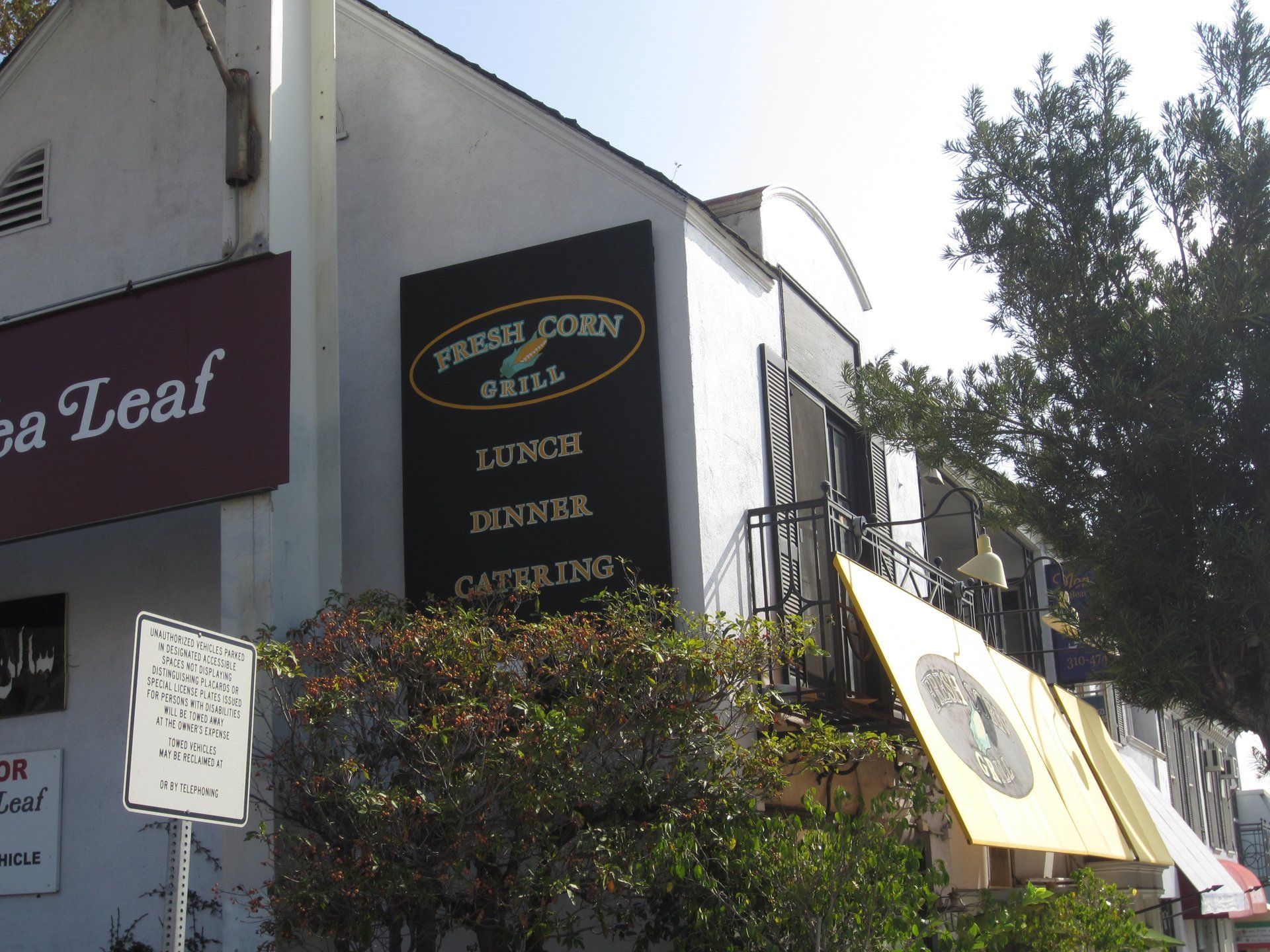 Street view of a corner building with café signs and outdoor awnings, partly shaded by trees.