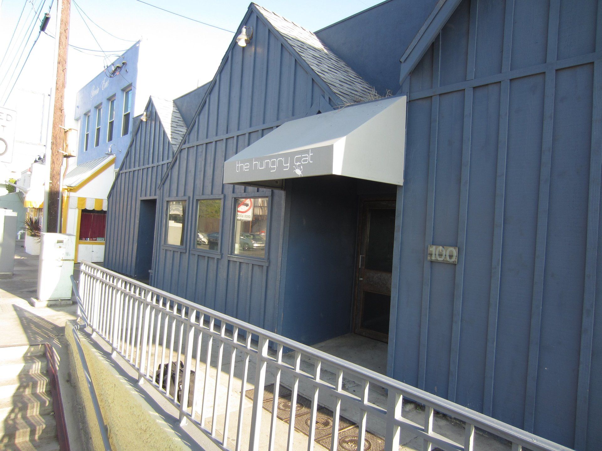 Blue-gray building with steep roofs, entrance awning, and metal railing along a sunny sidewalk.